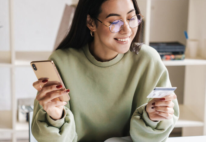 Young woman sits at desk while researching how to pay off credit card debt on her mobile phone.