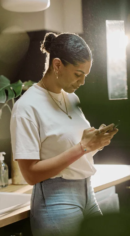 Woman in a white t-shirt looking at her phone in a warmly lit bathroom