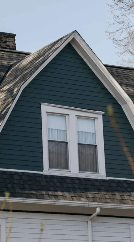 Upper gable of a house with dark green siding, white trim, double window, and a gray shingled mansard-style roof