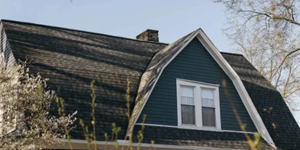 Upper gable of a house with dark green siding, white trim, double window, and a gray shingled mansard-style roof